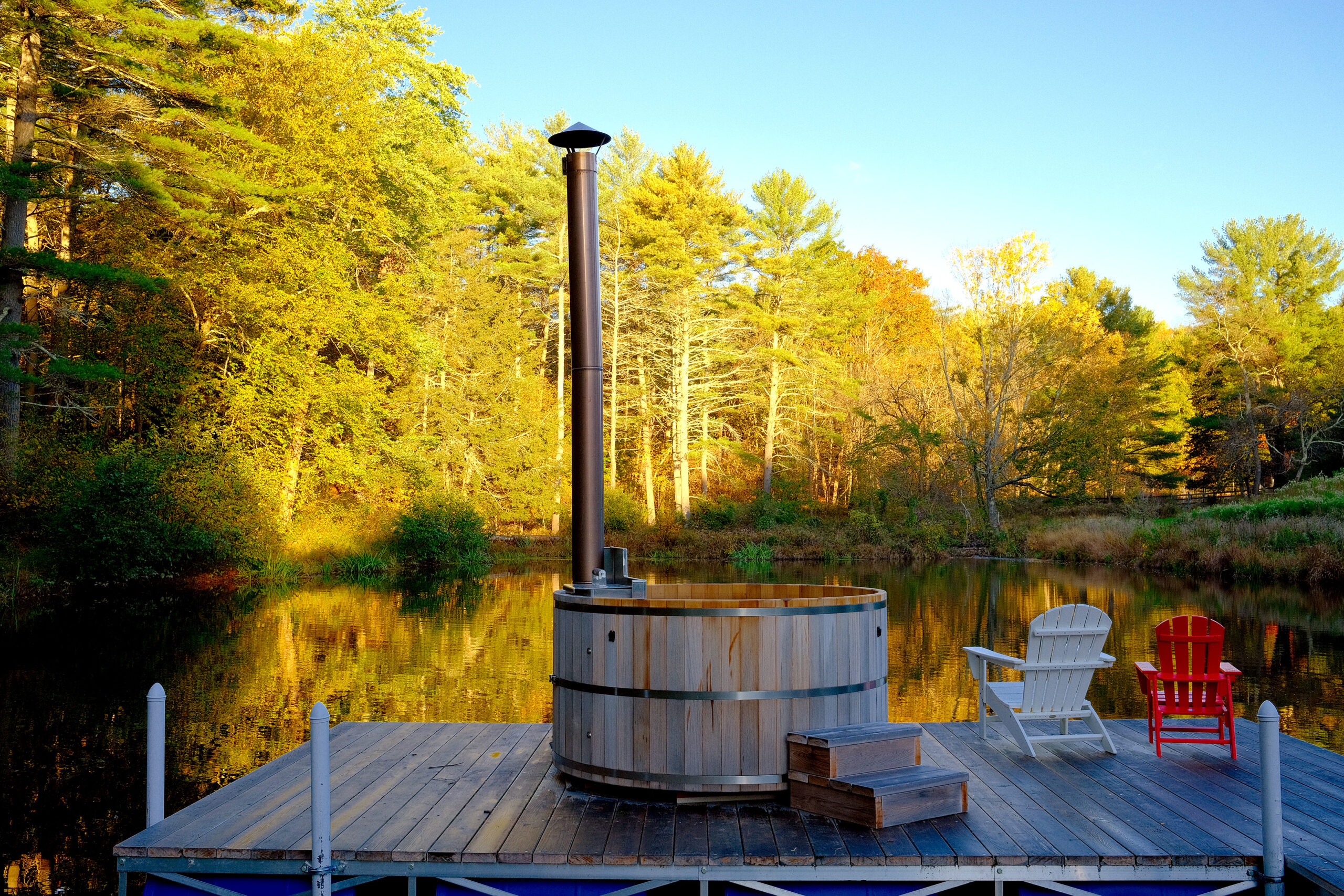 Wood-fired hot tub on a deck with two chairs next to it on a pond. 